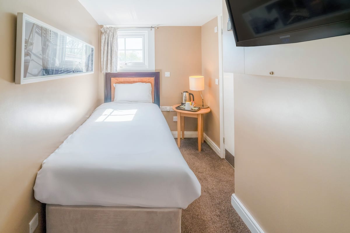 A small, neatly arranged hotel room with a single bed, a bedside table with a lamp, a television mounted on the wall, and a window providing natural light.