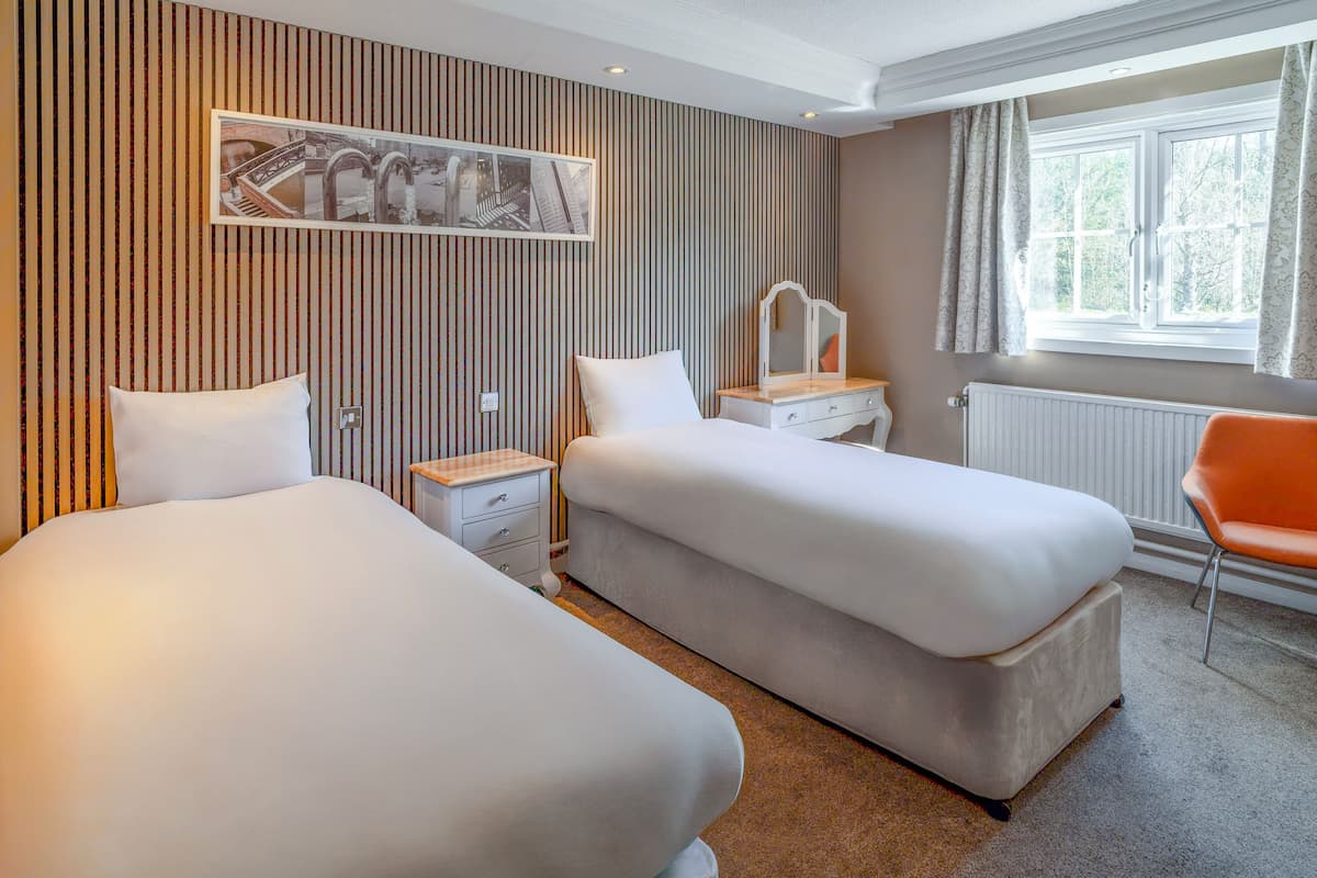 A hotel room with two beds, a bedside table with a mirror, a chair, and a window with curtains.