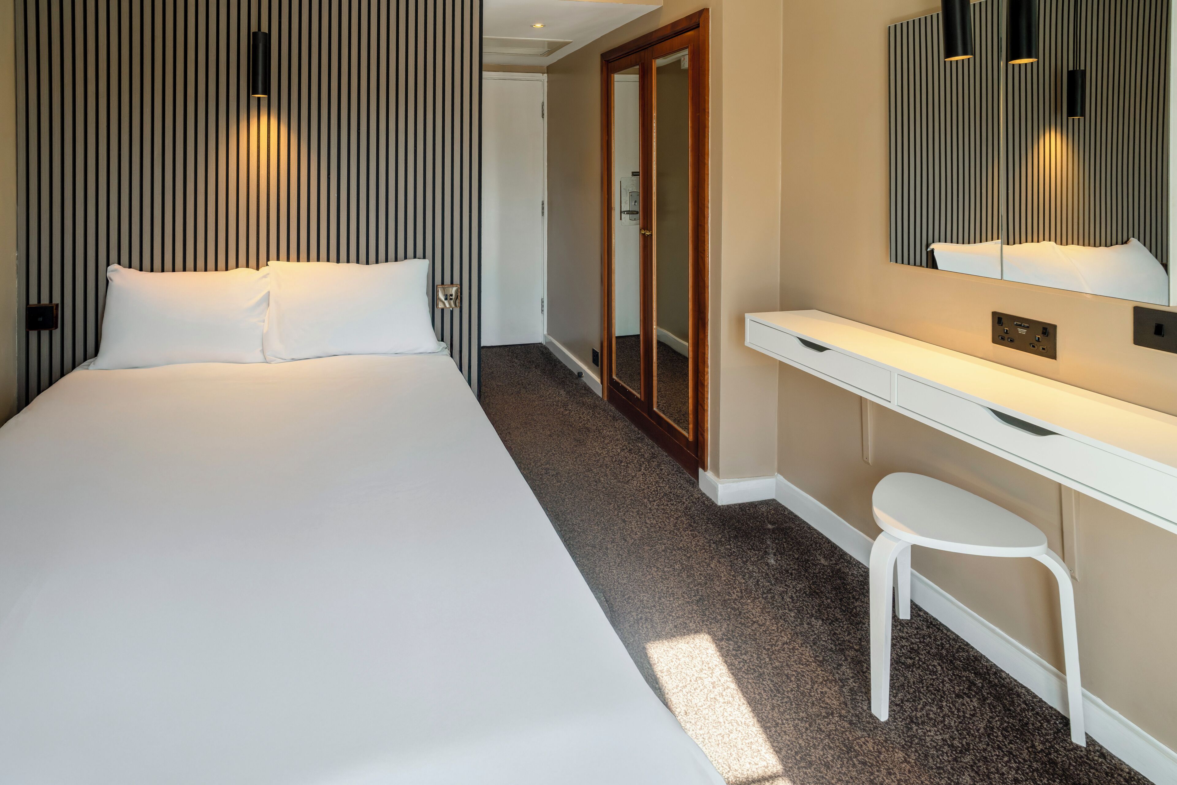 A hotel room with a large bed, a white desk, a small white stool, a mirror, and a wardrobe with a sliding door.