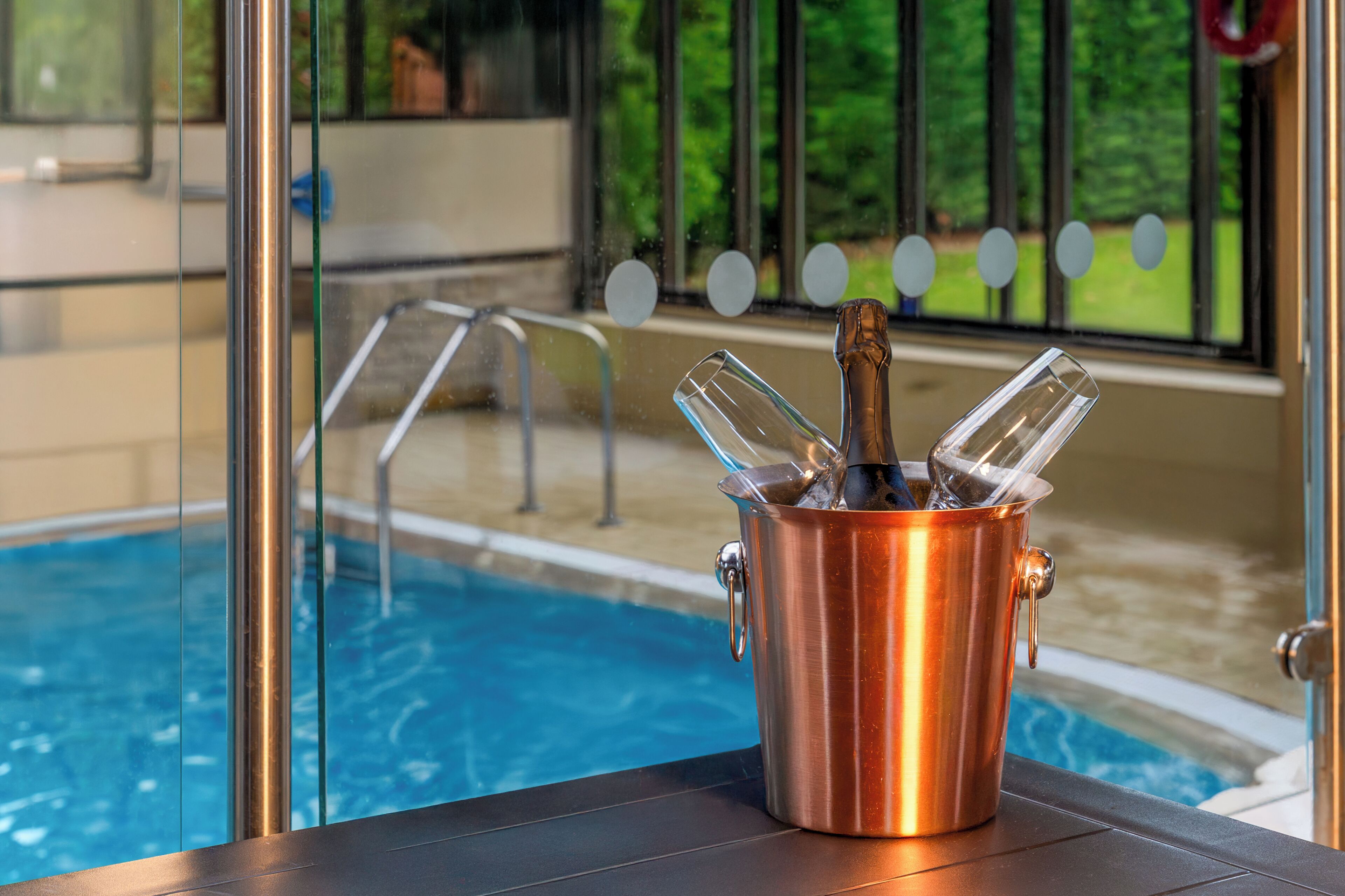 A copper bucket with a bottle and glasses on a poolside table, overlooking a swimming pool.