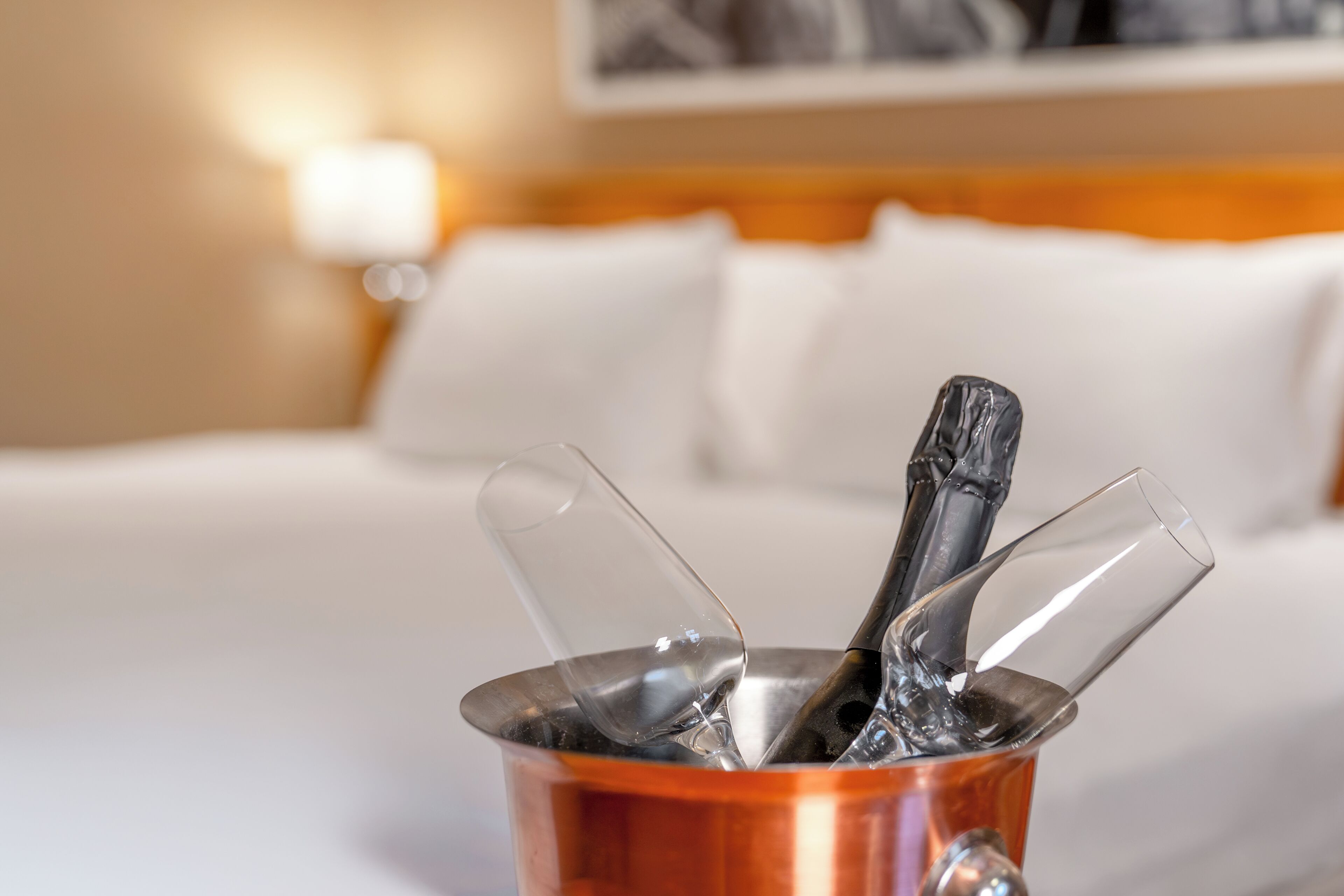 A hotel room with a bed, a nightstand, and a bucket containing a bottle and glasses.