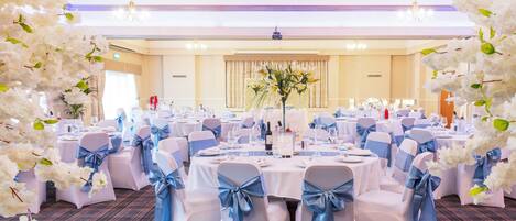 A banquet hall with round tables, white chairs with blue sashes, and white floral decorations.