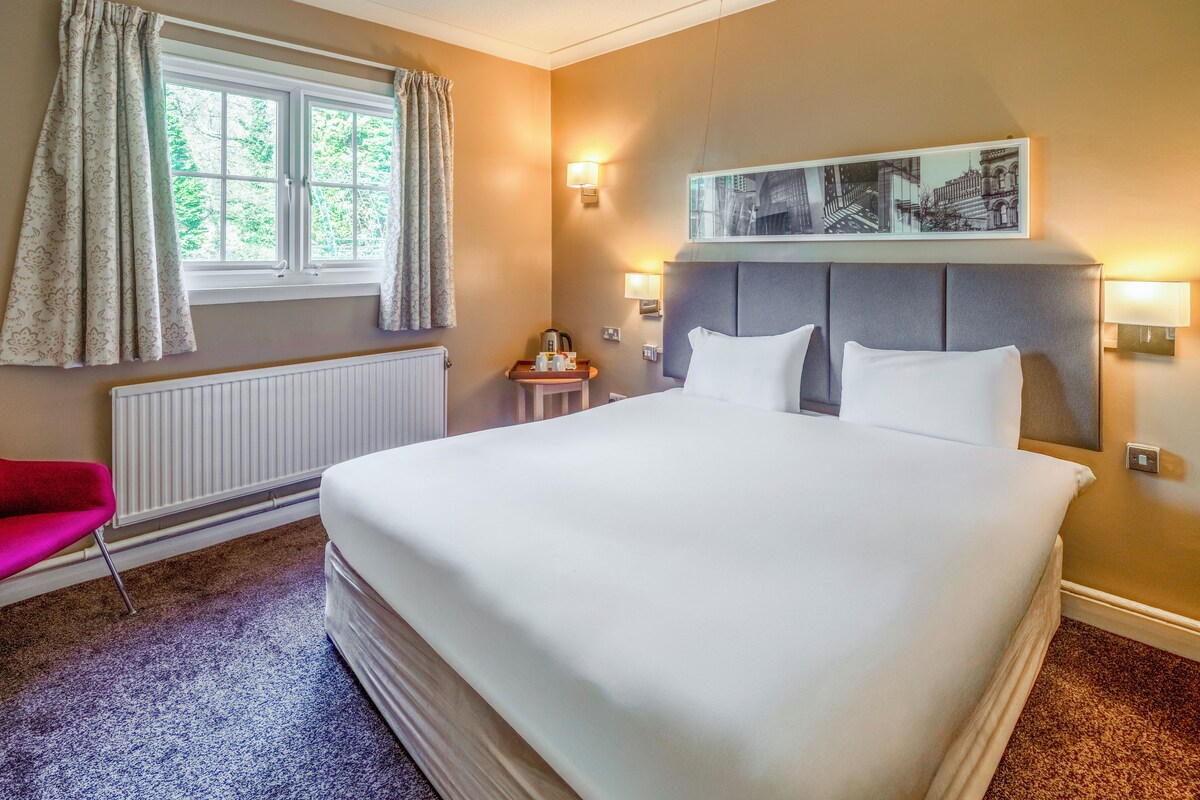 A hotel room with a large bed, a window with curtains, a radiator, a small table with a lamp, and a framed picture on the wall.