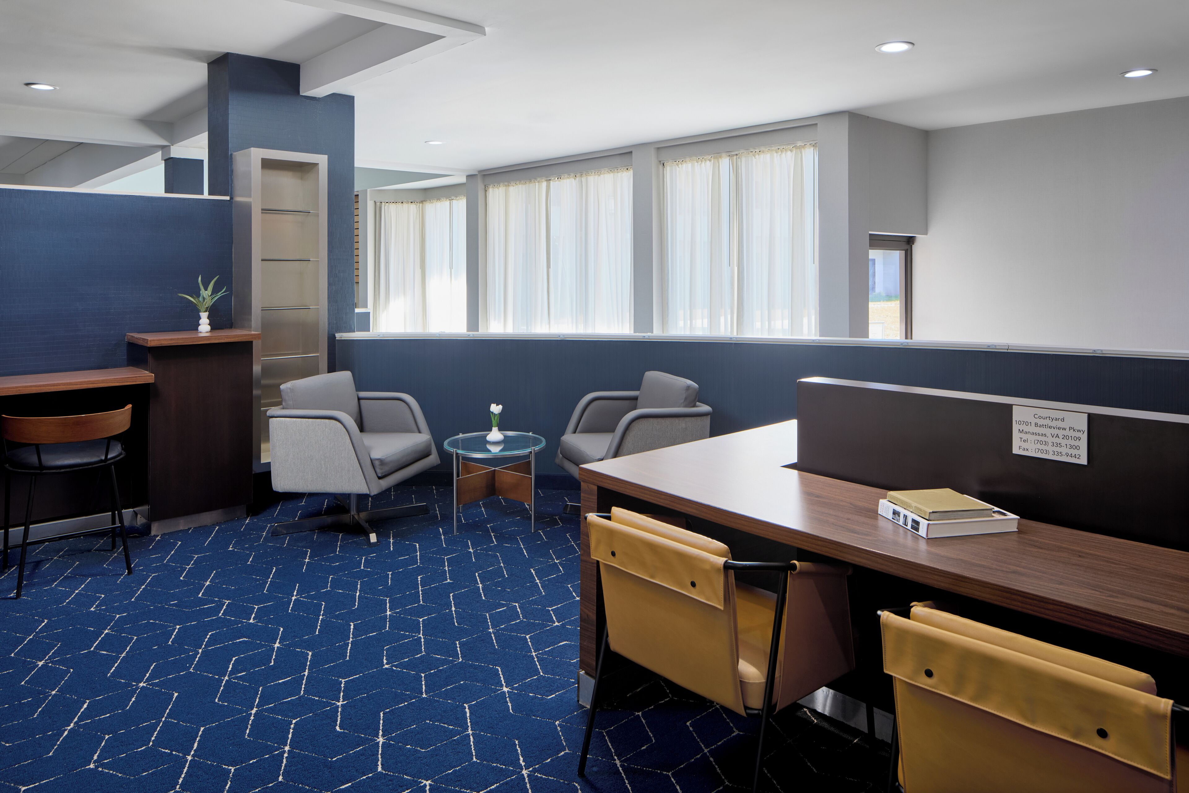 Une chambre d’hôtel avec un tapis à motifs bleus, un bureau en bois, deux chaises, une petite table avec un vase, et une vue sur l’extérieur grâce à de grandes fenêtres.