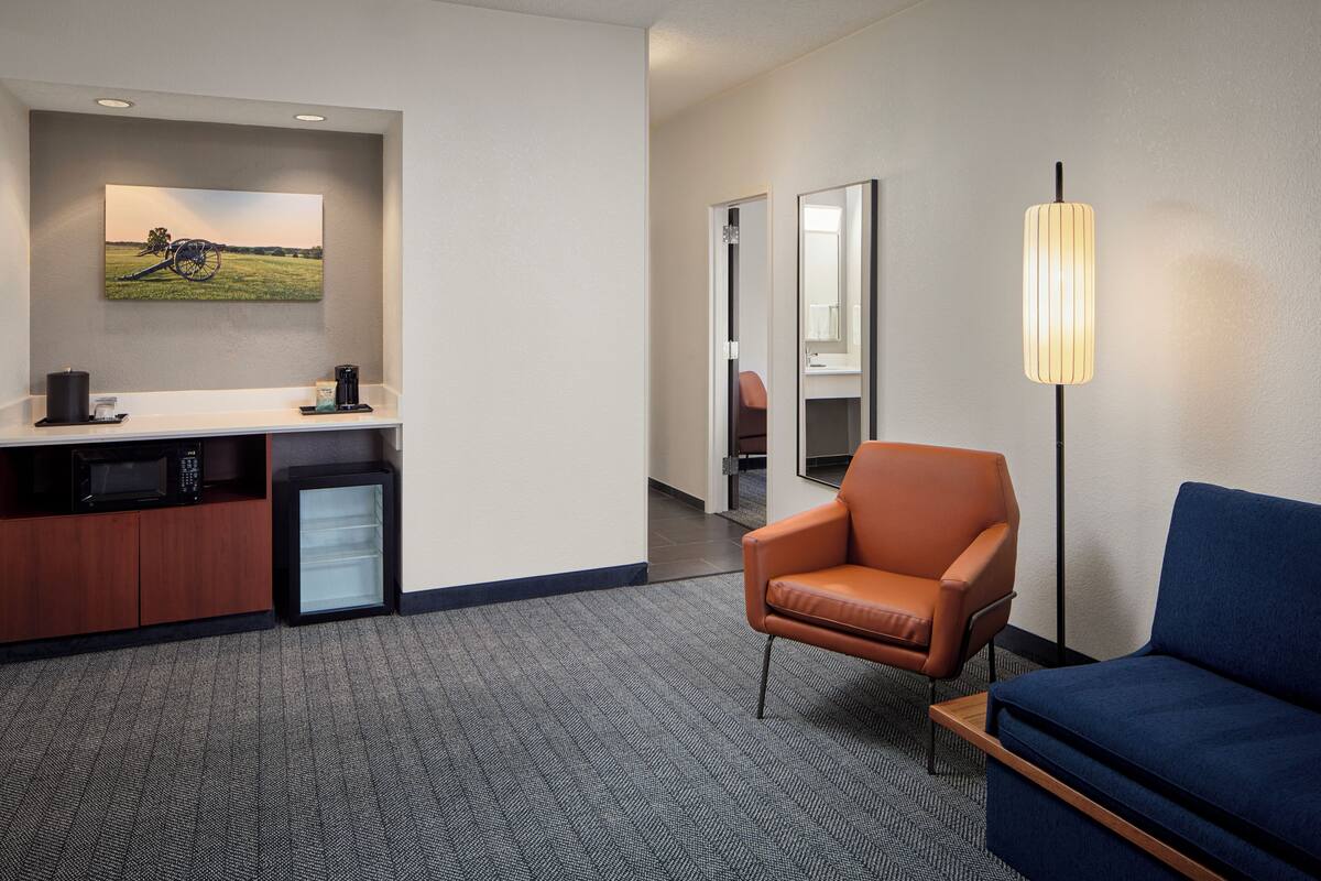 A hotel room with a kitchenette, a blue sofa, an orange armchair, a standing lamp, and a painting on the wall.