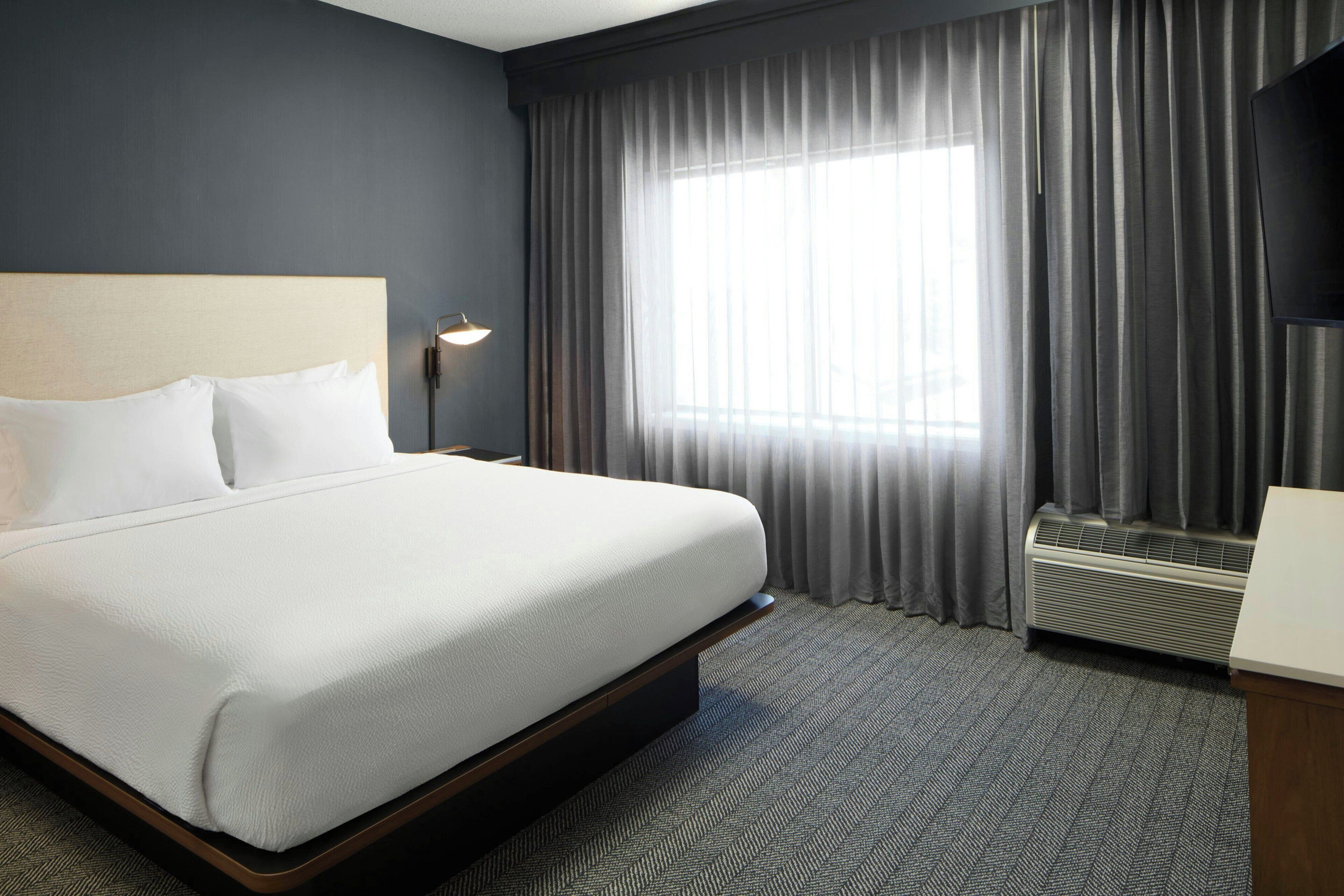 Una habitación de hotel con cama, mesita de noche, lámpara, ventana con cortinas y equipo de aire acondicionado.
