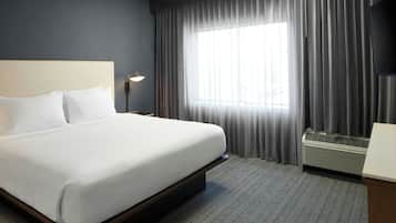 Una habitación de hotel con cama, mesita de noche, lámpara, ventana con cortinas y equipo de aire acondicionado.