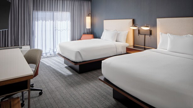 A hotel room with two beds, a desk, a chair, and a large window with curtains.