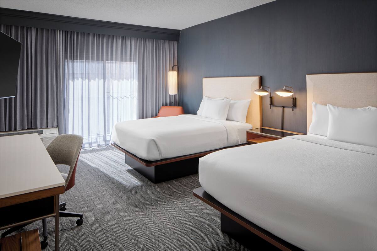 A hotel room with two beds, a desk, a chair, and a large window with curtains.