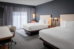 Habitación de hotel con dos camas, un escritorio, una silla y un ventanal con cortinas.