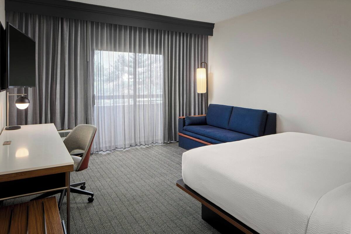 A hotel room with a bed, a desk, a chair, a TV, and a window with curtains.