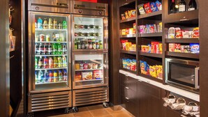 A kitchenette with a refrigerator stocked with beverages and snacks, a microwave, and shelves with packaged food items.