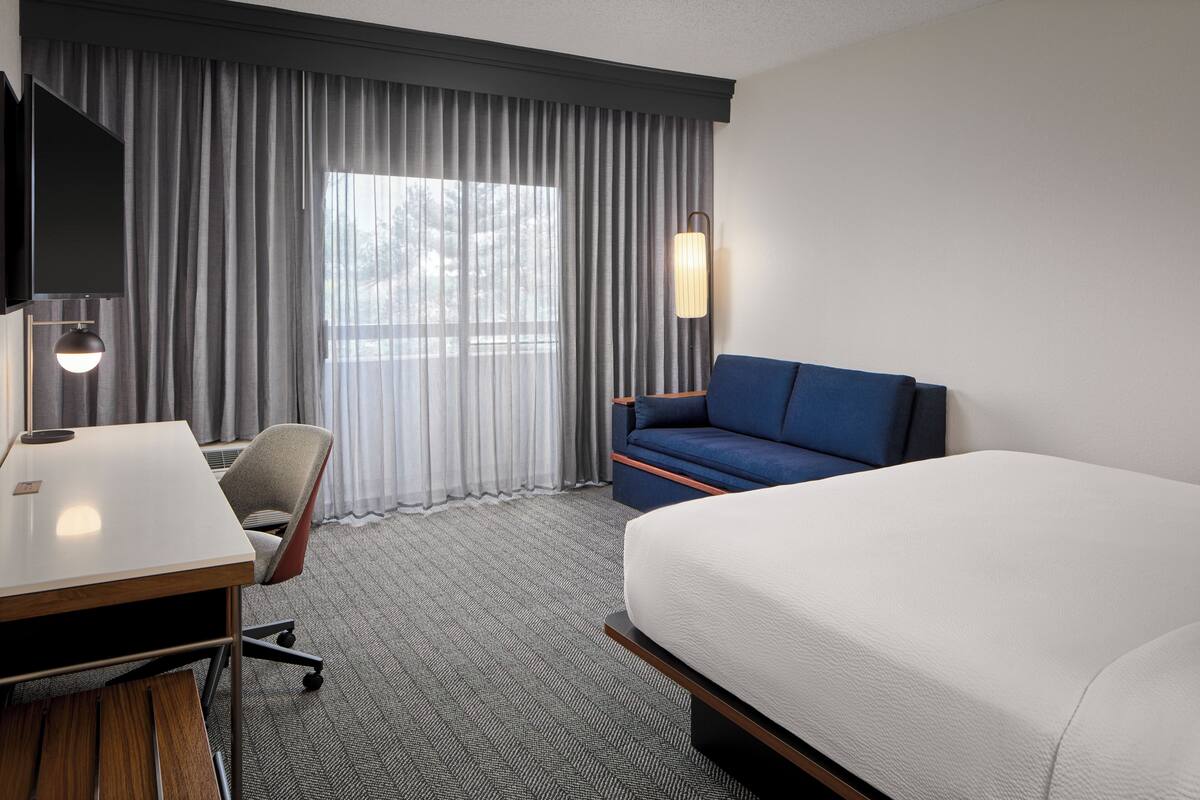A hotel room with a bed, a desk, a chair, a TV, and a window with curtains.
