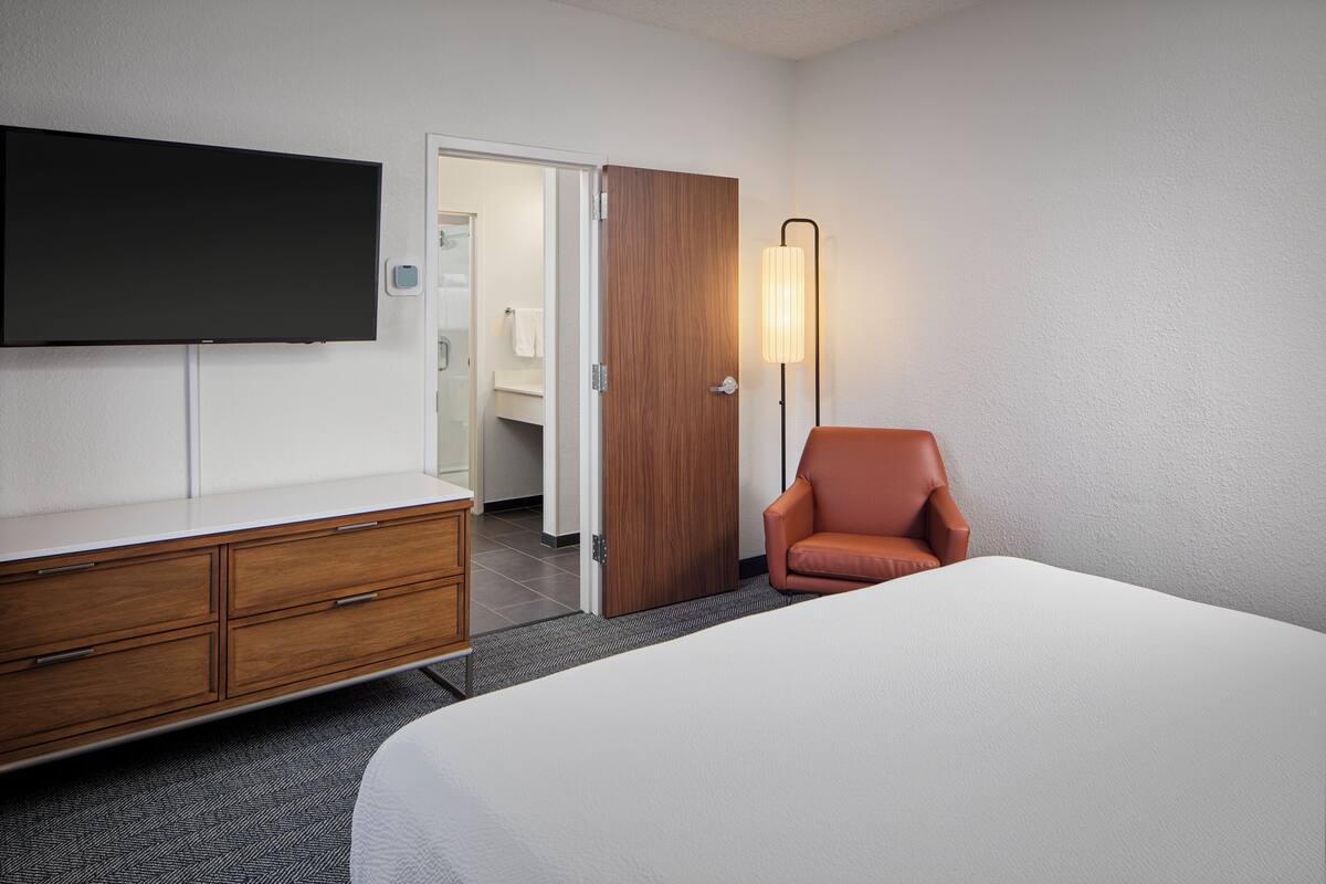 Ein Hotelzimmer mit einem Bett, einem an der Wand montierten Fernseher, einer Holzkommode, einem Ledersessel und einer Tür zu einem Badezimmer.