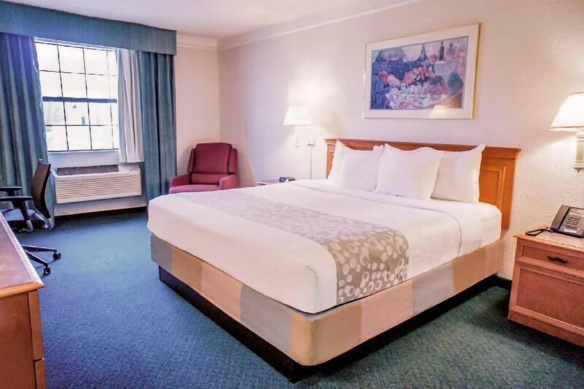 A hotel room with a large bed, a desk, a chair, a window with curtains, a painting on the wall, and a nightstand with a lamp.