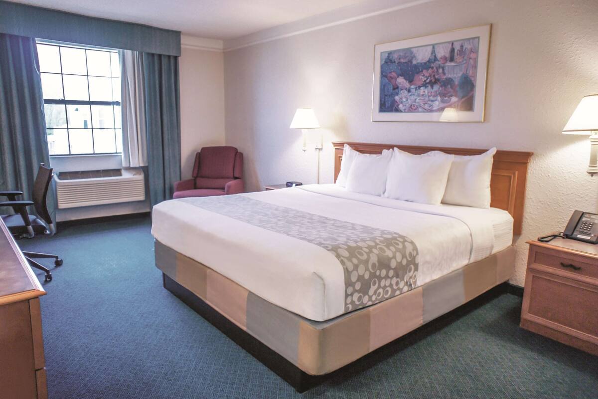 A hotel room with a large bed, a desk, a chair, a radiator, and a painting on the wall.
