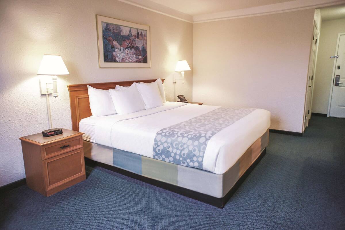 A hotel room with a bed, bedside lamps, a nightstand, a painting on the wall, and a door.