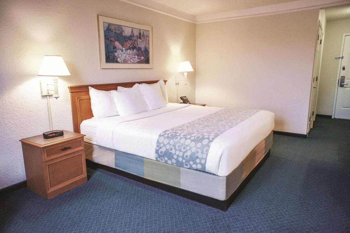 A hotel room with a bed, bedside lamps, a nightstand, a painting on the wall, and a door to the right.