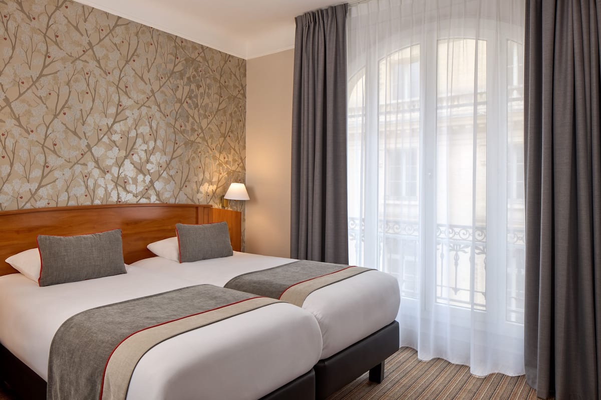 Habitación de hotel con dos camas, un ventanal grande con cortinas, un papel tapiz estampado y un cabecero de madera.