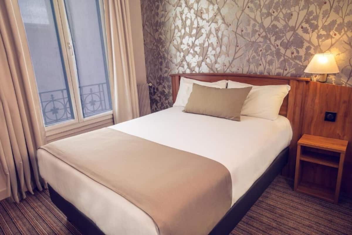 Una habitación de hotel con una cama grande, una mesita de noche con lámpara, una ventana con cortinas y una pared con estampado floral.