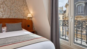 Une chambre d’hôtel avec un grand lit, une tête de lit en bois, une table de chevet, une lampe et une vue sur la ville par une fenêtre donnant sur un balcon.