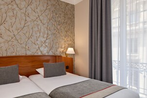 A hotel room with two beds, a wooden headboard, a wall with a floral pattern, a lamp, and a view of a balcony.