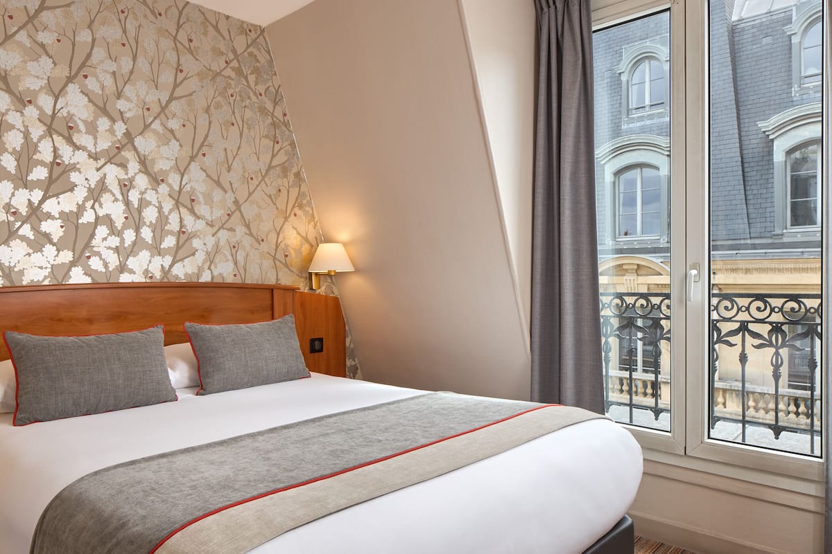Una cama bien hecha con una colcha gris y blanca, un cabecero de madera y dos almohadas grises. El papel tapiz con un diseño adorna la pared detrás de la cama.