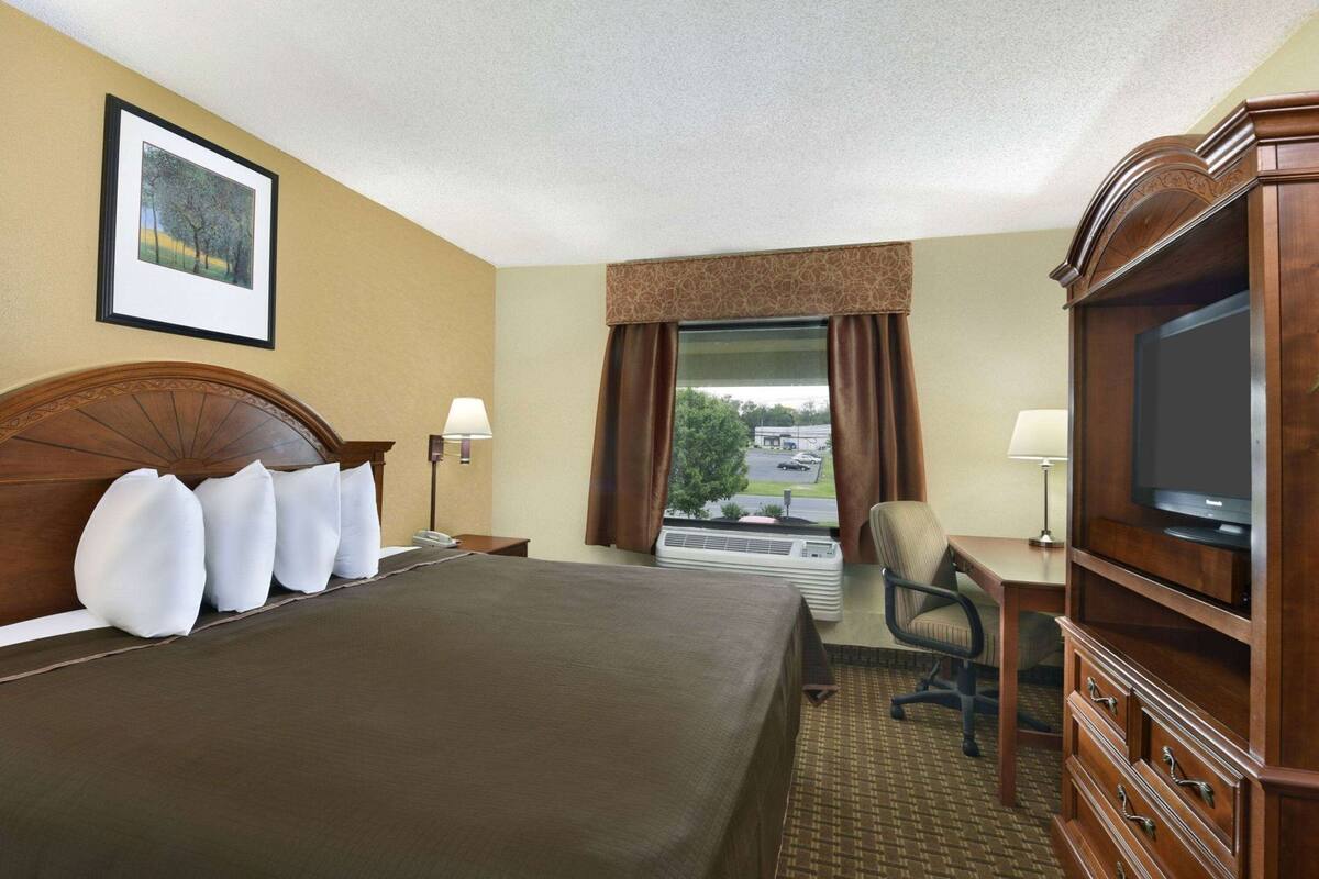Ein Hotelzimmer mit einem großen Bett, einem Schreibtisch mit Fernseher, einem Sessel, einem Fenster mit Vorhängen und einem Bild an der Wand.