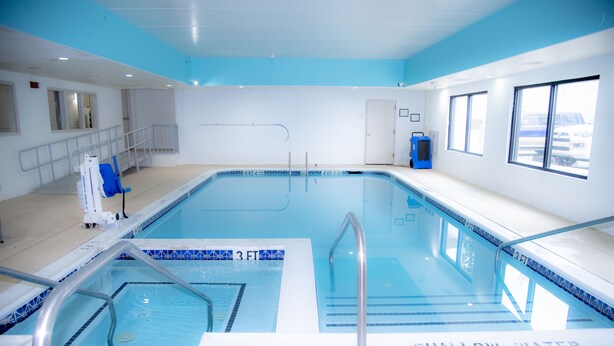 Una piscina con zona de aguas poco profundas señalizada, un vestuario con una toalla azul, y una ventana que da a coches aparcados.