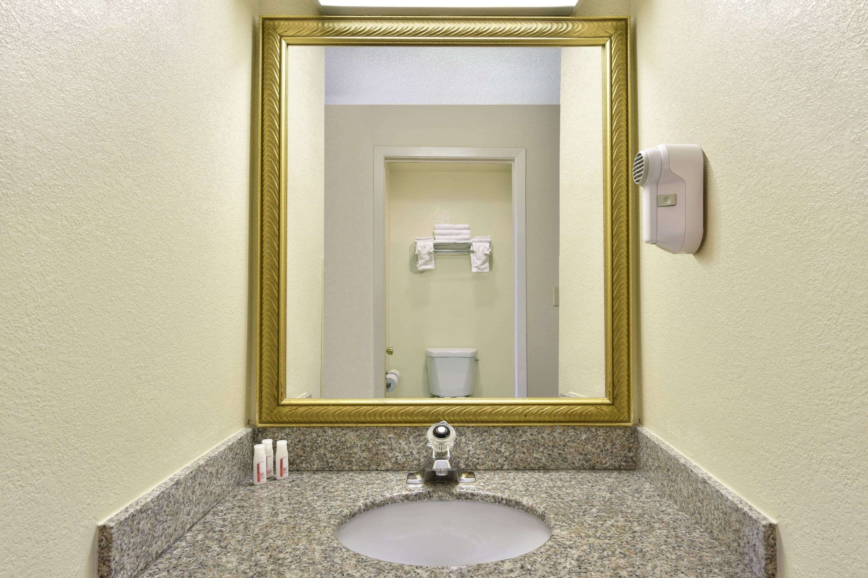 Un baño con lavamanos de granito, un espejo con marco dorado y un secamanos.