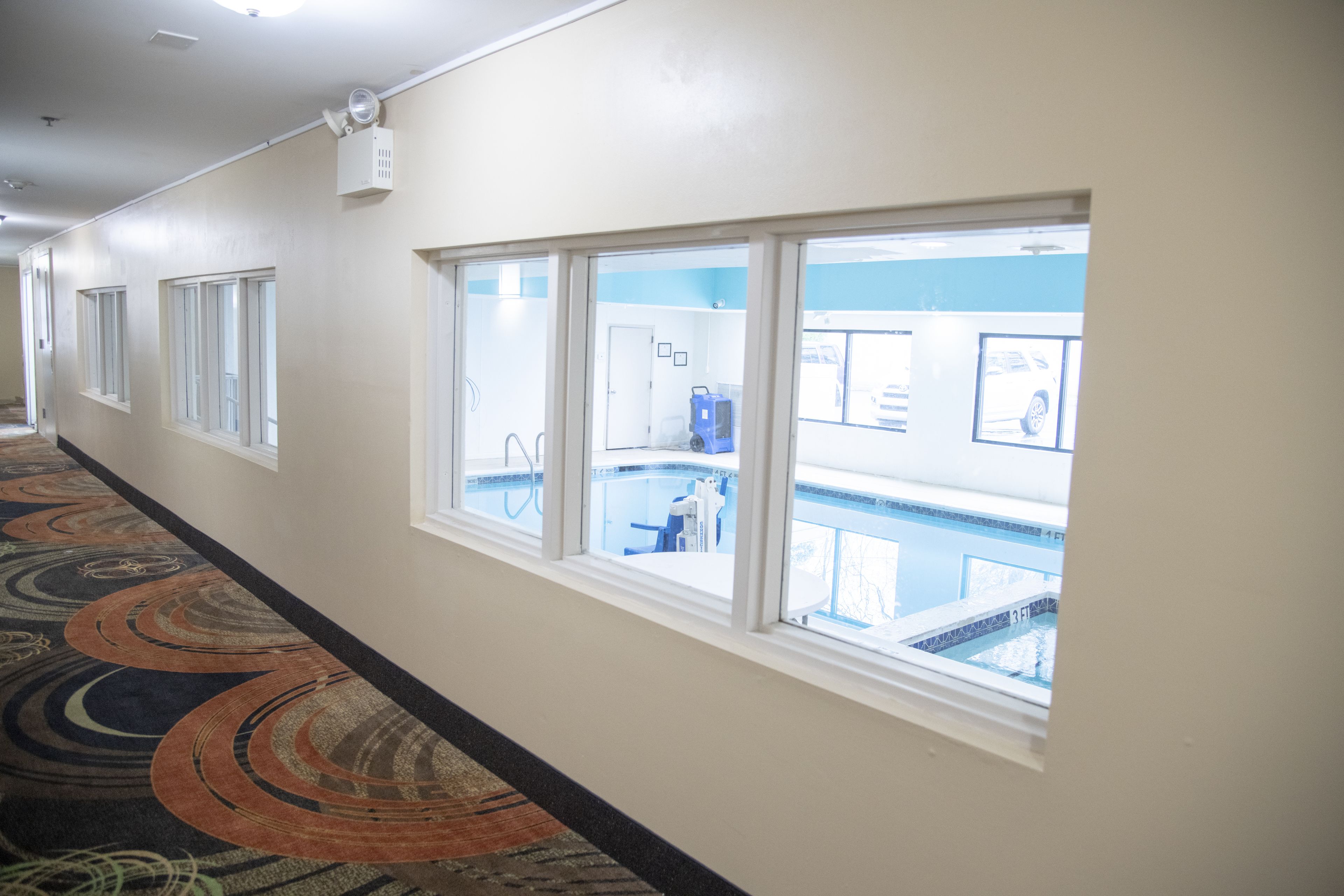 A hotel corridor with large windows, a carpeted floor, and a visible pool area.