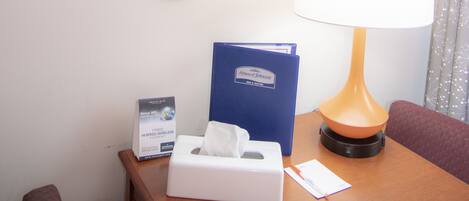 Habitación de hotel con una mesa de madera, una lámpara, una caja de pañuelos, una carpeta azul y varios espejos en la pared.