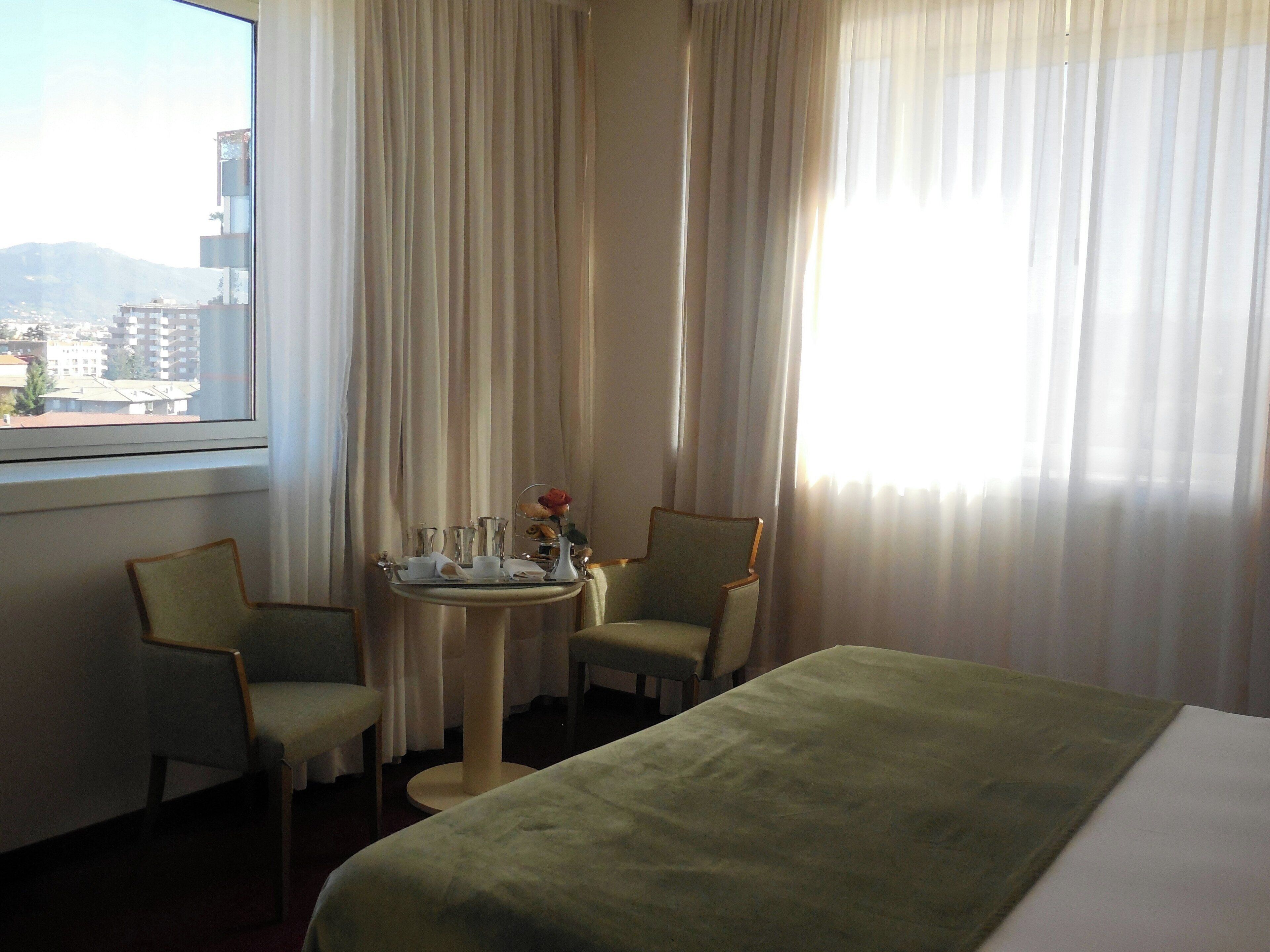 Habitación de hotel con cama, dos sillas, una mesita con una bandeja de desayuno y una ventana con cortinas.
