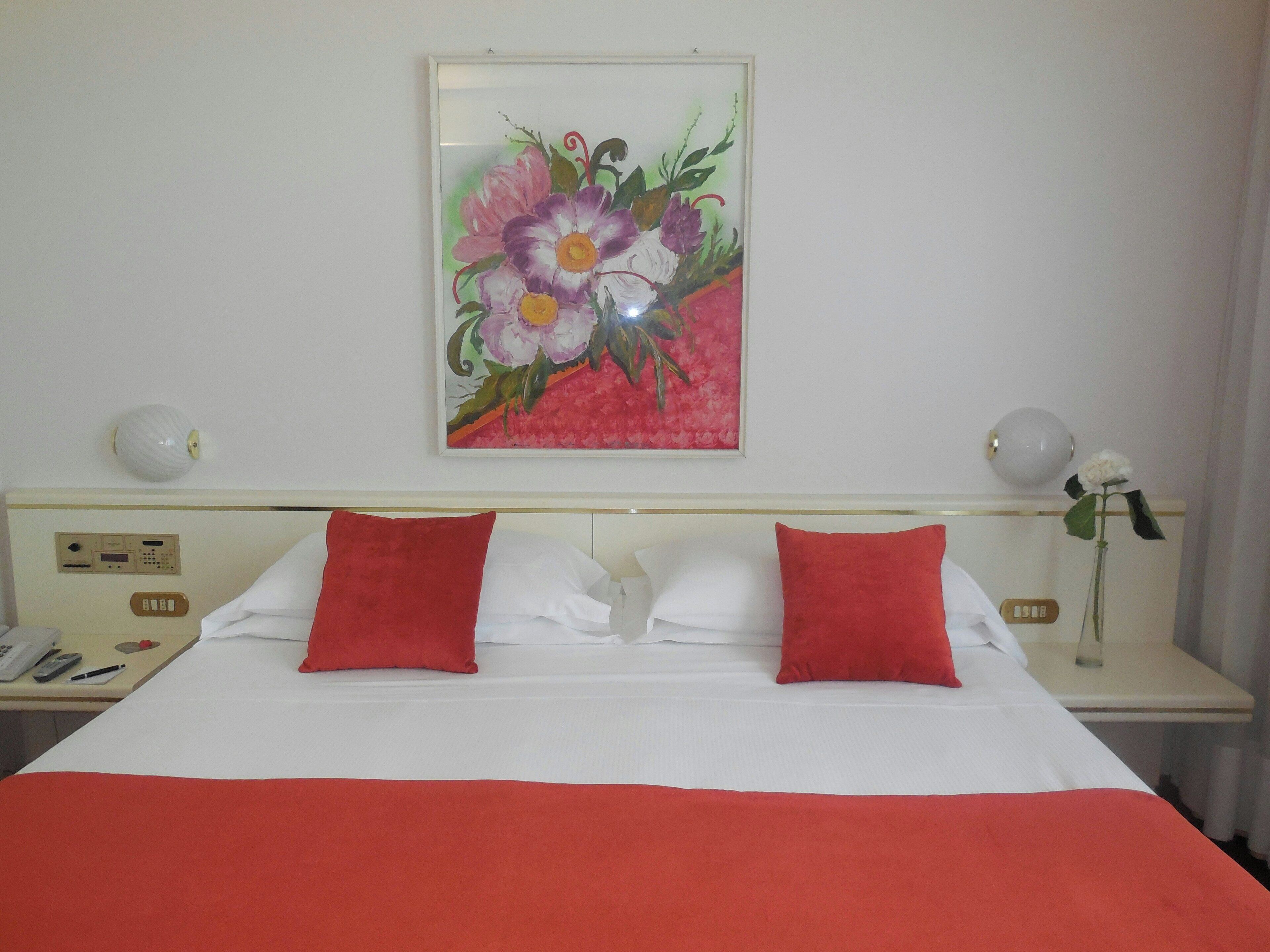 A hotel room with a bed, two red pillows, a white bedspread, a bedside table with a lamp, a phone, and a wall-mounted painting of flowers.