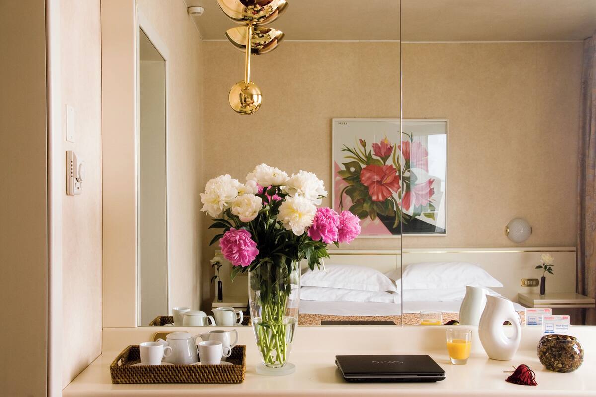 Une chambre d’hôtel avec un lit, une table de chevet ornée d’un vase de fleurs, un miroir et un plateau de petit-déjeuner.