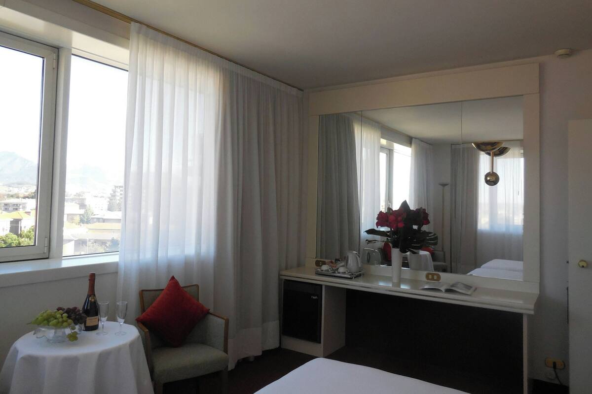 A hotel room with a bed, a desk with a mirror, a chair, a small table with a bottle and glasses, and a window with curtains.