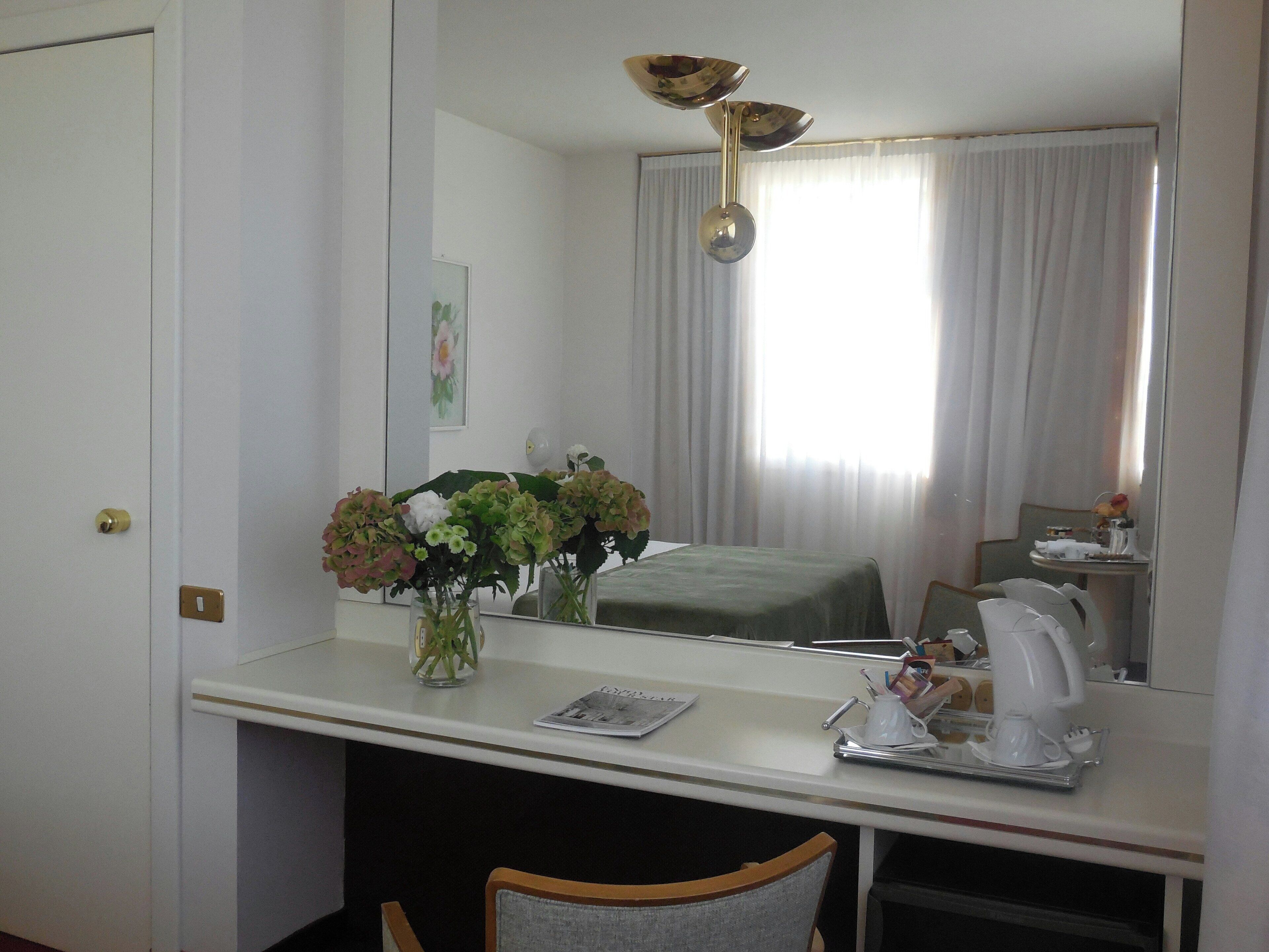 Hotelkamer met een wit bureau, een stoel, een bed met een groene sprei, een vaas met bloemen, een spiegel en een raam met gordijnen.