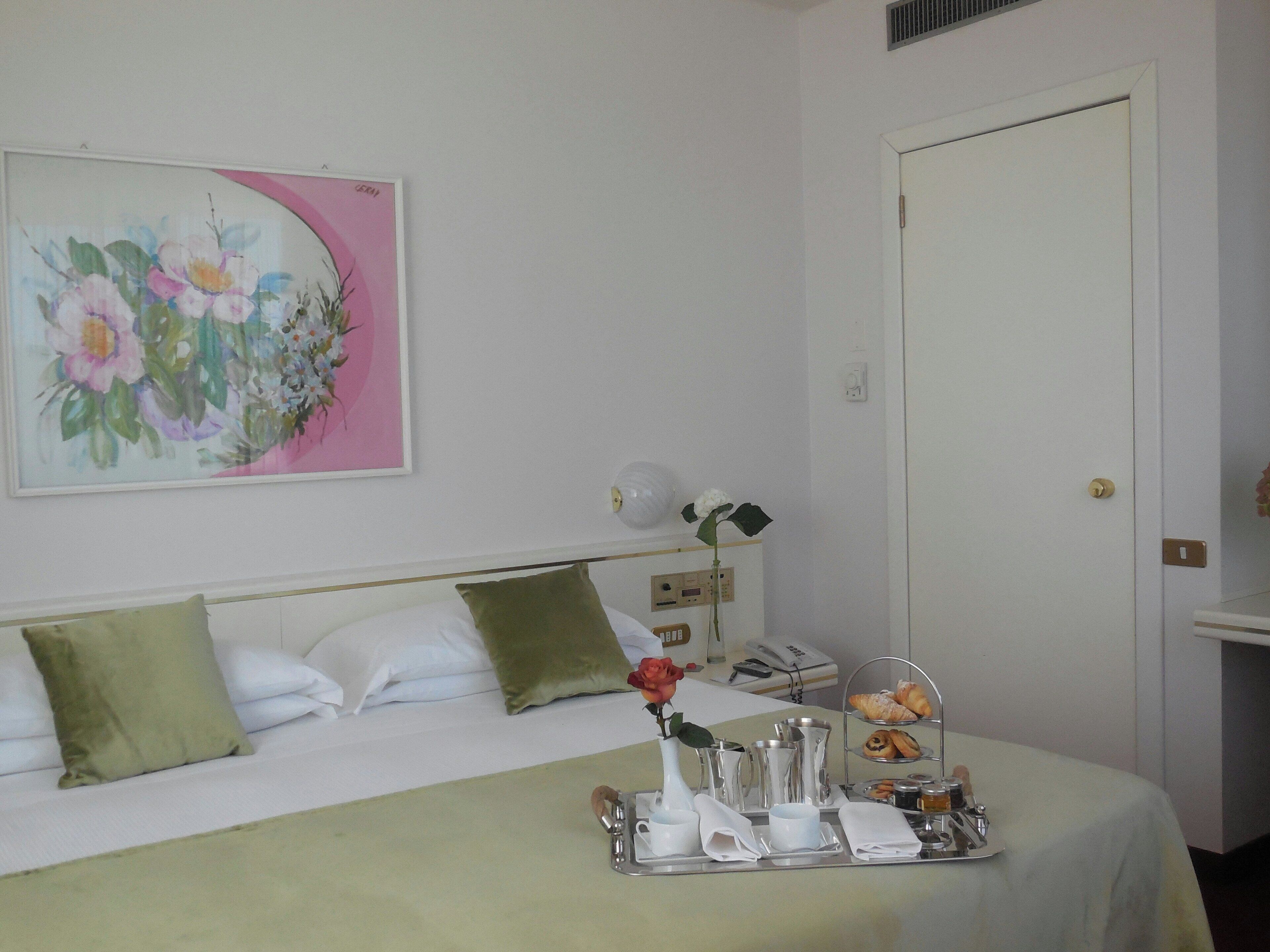 Una habitación de hotel con cama, almohadas, una bandeja con productos para el desayuno, un jarrón con una flor y un cuadro en la pared.