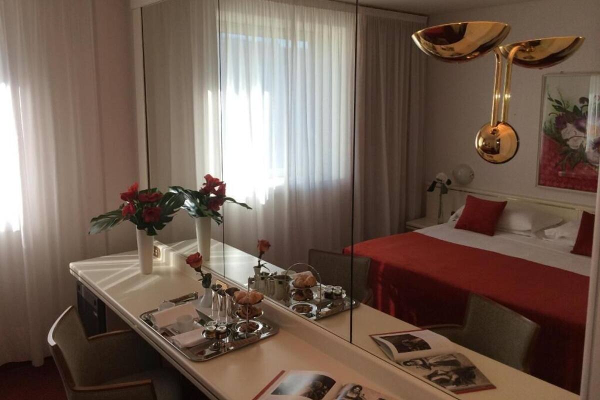 A hotel room with a bed, a dining table with a tray of food, a vase with flowers, and a large mirror.