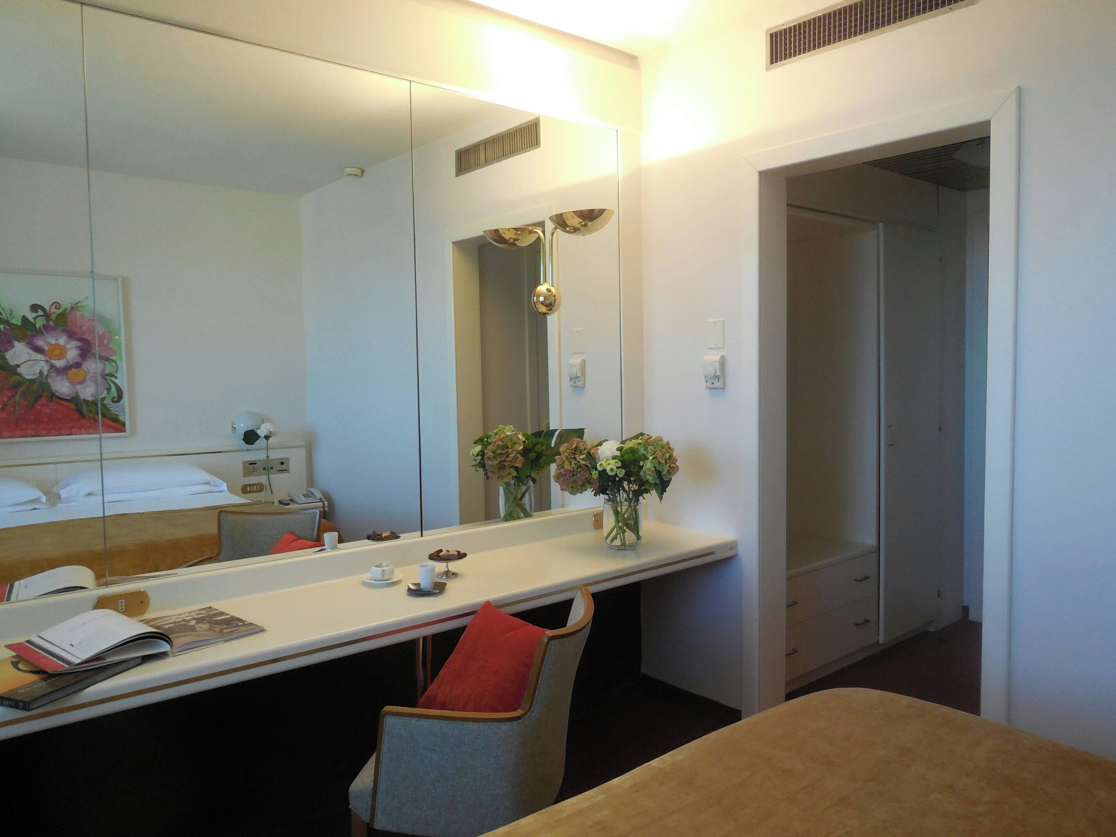 Una camera d'albergo con un letto, una scrivania con una sedia, uno specchio, un vaso con fiori e una porta che dà su un'altra stanza.