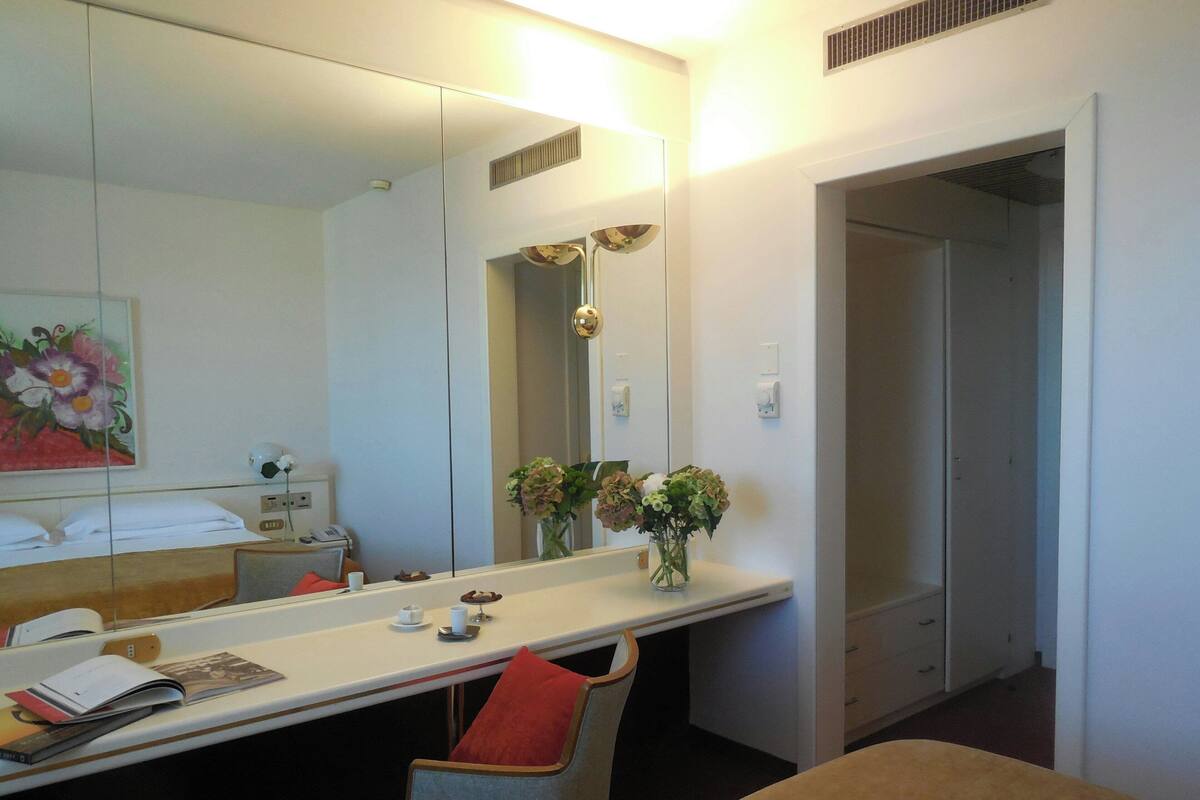 Une chambre d’hôtel comprenant un lit, un bureau avec une chaise, un miroir, un vase avec des fleurs et une porte donnant sur une autre pièce.