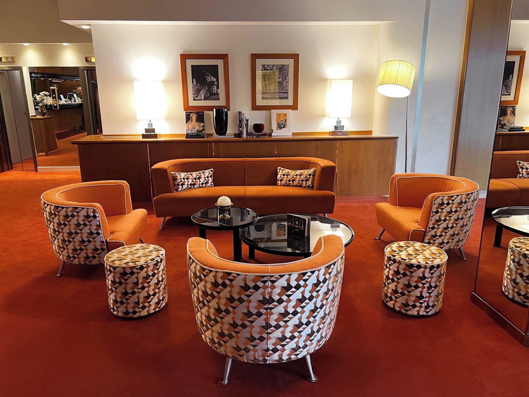 A hotel lobby with a reception desk, a sofa, patterned armchairs, ottomans, and framed artwork on the wall.