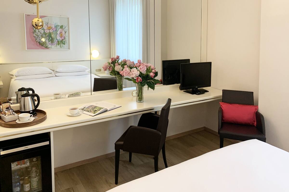 A hotel room with a bed, a desk with a computer, a chair, a coffee maker, and a vase with flowers.