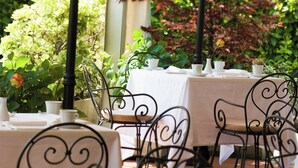 An outdoor dining area with tables set for four, surrounded by greenery and red foliage.