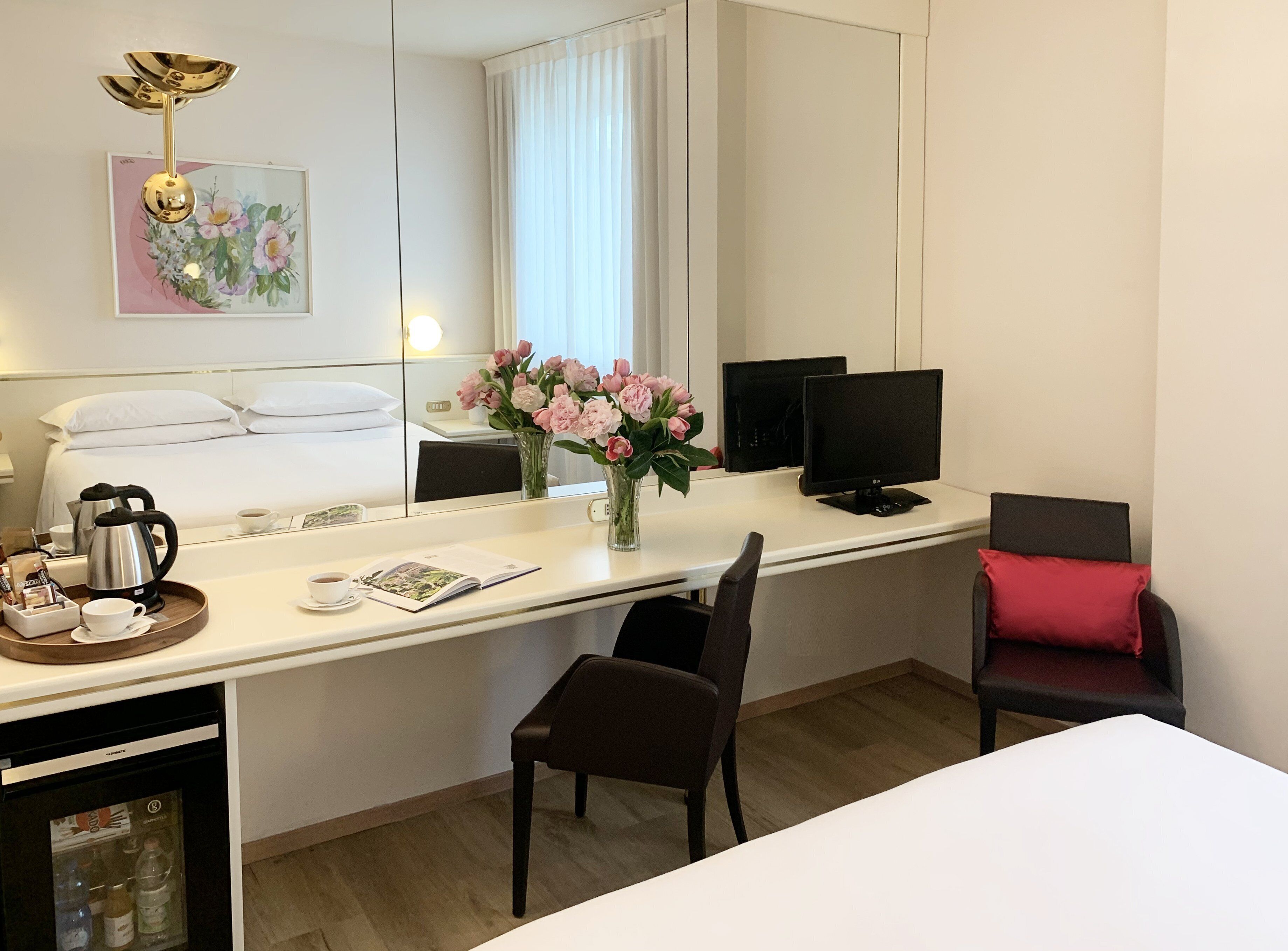 Habitación de hotel con una cama, un escritorio con computadora, una silla, una cafetera y un jarrón con flores.
