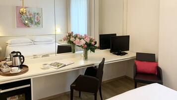 Habitación de hotel con una cama, un escritorio con computadora, una silla, una cafetera y un jarrón con flores.