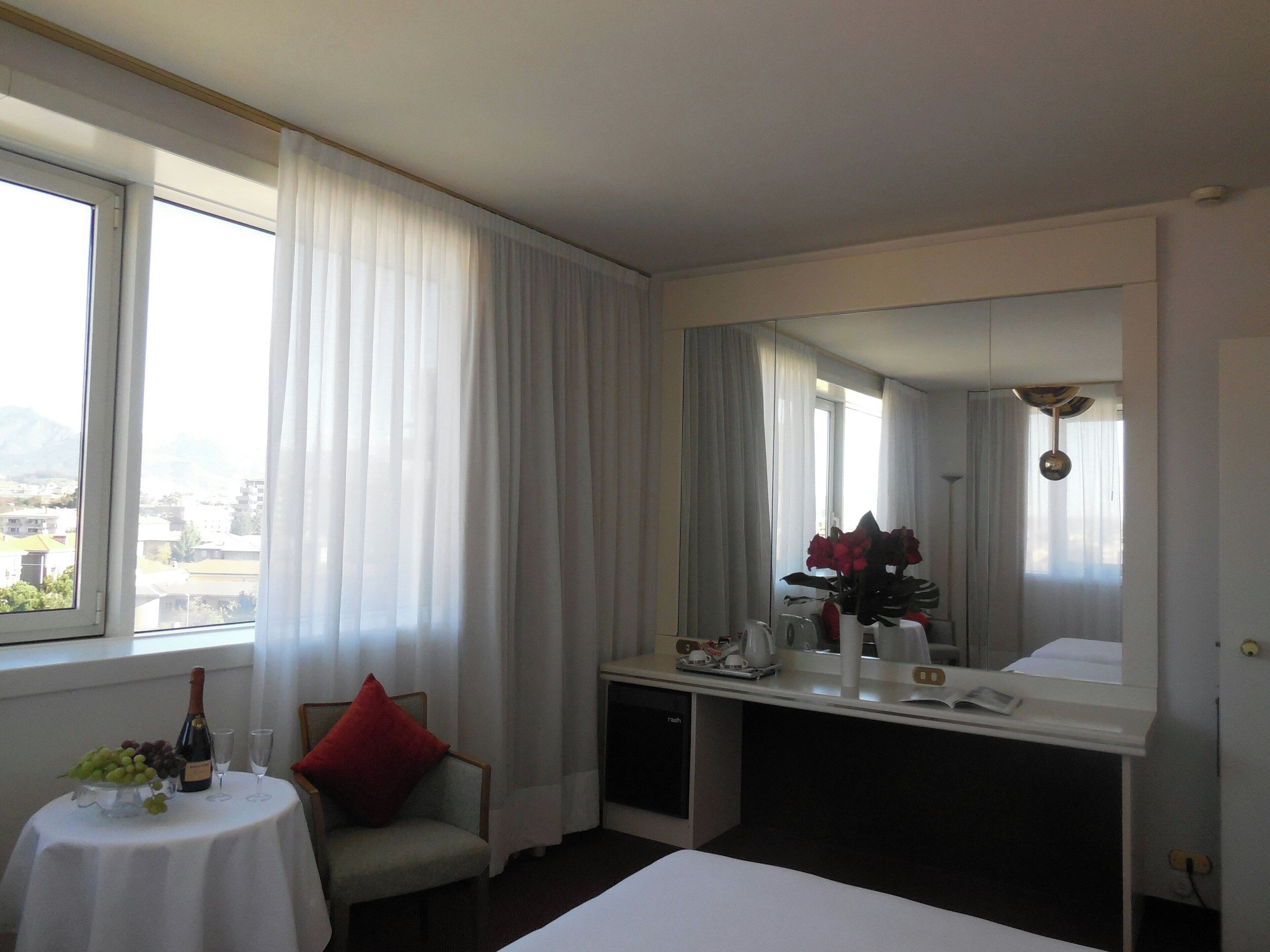 A hotel room with a bed, a desk with a mirror, a chair, a small table with a bottle and glasses, and a window with curtains.