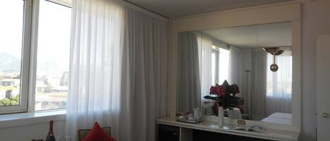 A hotel room with a bed, a desk with a mirror, a chair, a small table with a bottle and glasses, and a window with curtains.