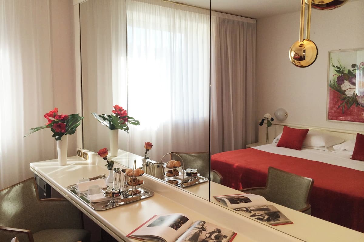 A hotel room with a large mirror, a bed with red bedding, a dining table with a tray of pastries, and a vase with red flowers.