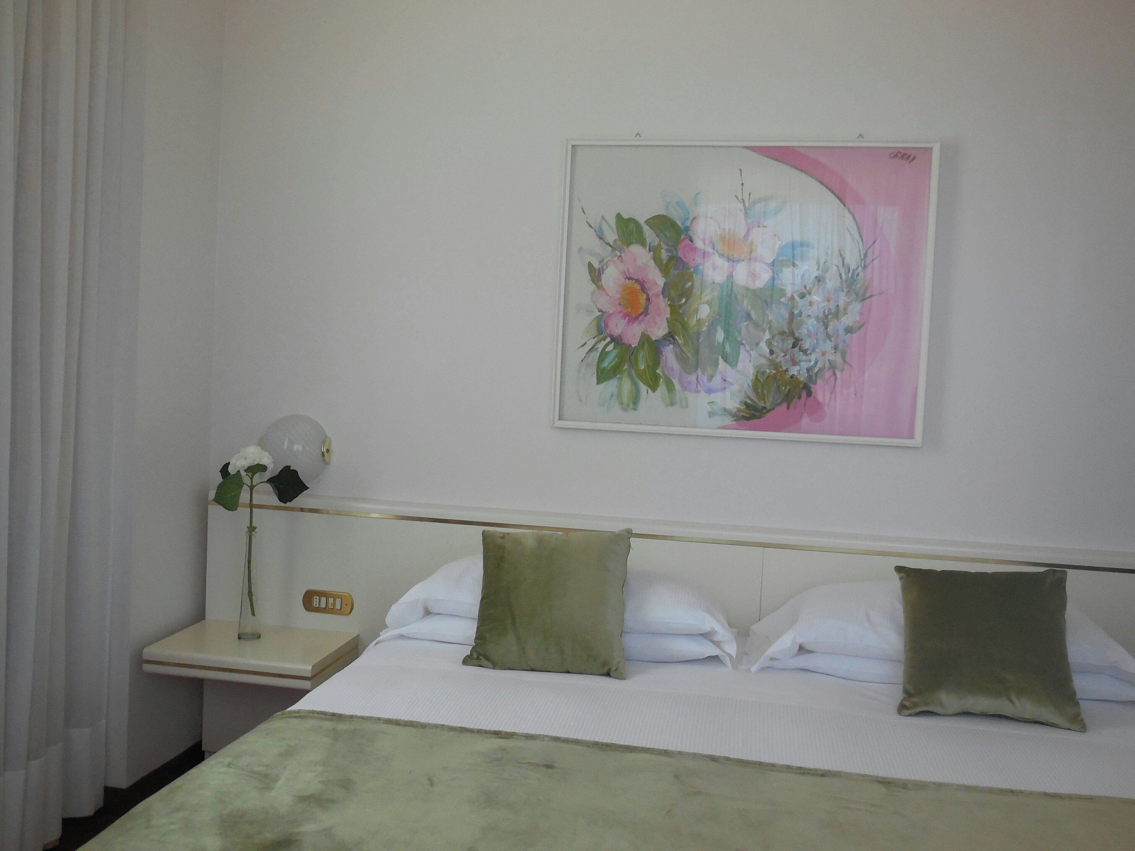 Een hotelkamer met een bed, kussens, een nachtkastje, een lamp en een bloemstil afbeelding aan de muur.
